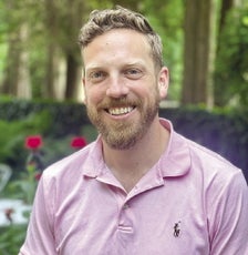 A man with blonde hair and a blonde beard wears a pink polo shirt in front of a garden.