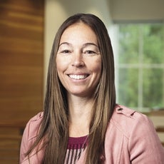 A woman with long brown hair wears a light pink suede jacket and a purple, pink, and black patterned sweater.