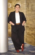 A woman with brown shoes, a dark suit, and a white shirt leans against a column.