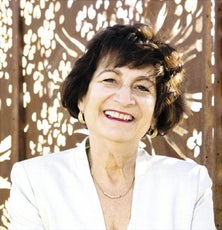 A headshot of a woman with short brown hair and bangs wears red lipstick and a white blazer