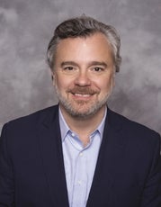 A headshot of a man with white and grey hair and beard wears a dark blue suit jacket and light blue button down
