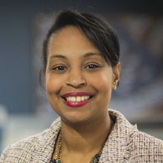 A headshot of a woman with dark hair pulled back and hangs wears a tweed suit jacket and red lipstick