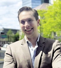 A man stands with his arms folded in front a city street while wearing a tan suit jacket and white button down