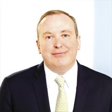 A man with short blonde hair wears a black suit jacket, white button down, and gold tie.