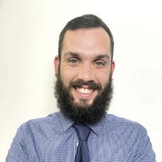 A man with dark hair and a beard wears a blue button down and tie.