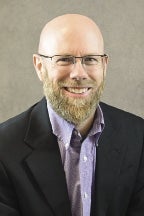 A bald man with a blonde beard wears a black blazer and purple button down.