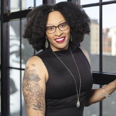 A woman with black curly hair wears a black mock-neck tank top.