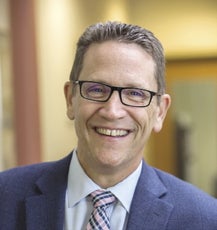 A man with short light brown hair wears black-rimmed glasses, a blue suit jacket, white button down, and pink/blue/white plaid tie.