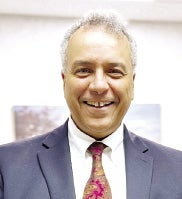 A man with a dark blue suit jacket, white button down, and brown and purple tie smiles for the camera.