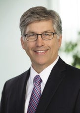 A man with gray hair wears a dark suit with a purple tie