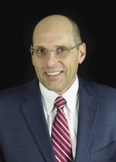 A headshot of a man wearing rimless glasses, a dark blue suit jacket, white button down, and a red and white striped tie.