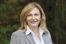 A woman with blonde hair wears a grey suit jacket, a white button down, and a pearl necklace.