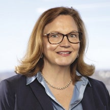 A woman with shoulder length blonde hair wears glasses, a black suit jacket, and light blue silk button down.