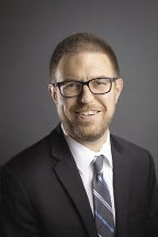 A headshot photo of a man in a dark suit with a blue tie.