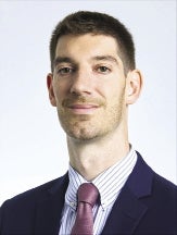A headshot of a man wearing a dark blue suit jacket, a striped button down, and a purple tie