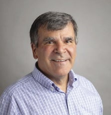 A man with short grey hair wears a blue and white striped button down.
