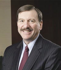 A man with short brown hair and a mustache wears a pinstripe black suit jacket, white button down, and red tie.