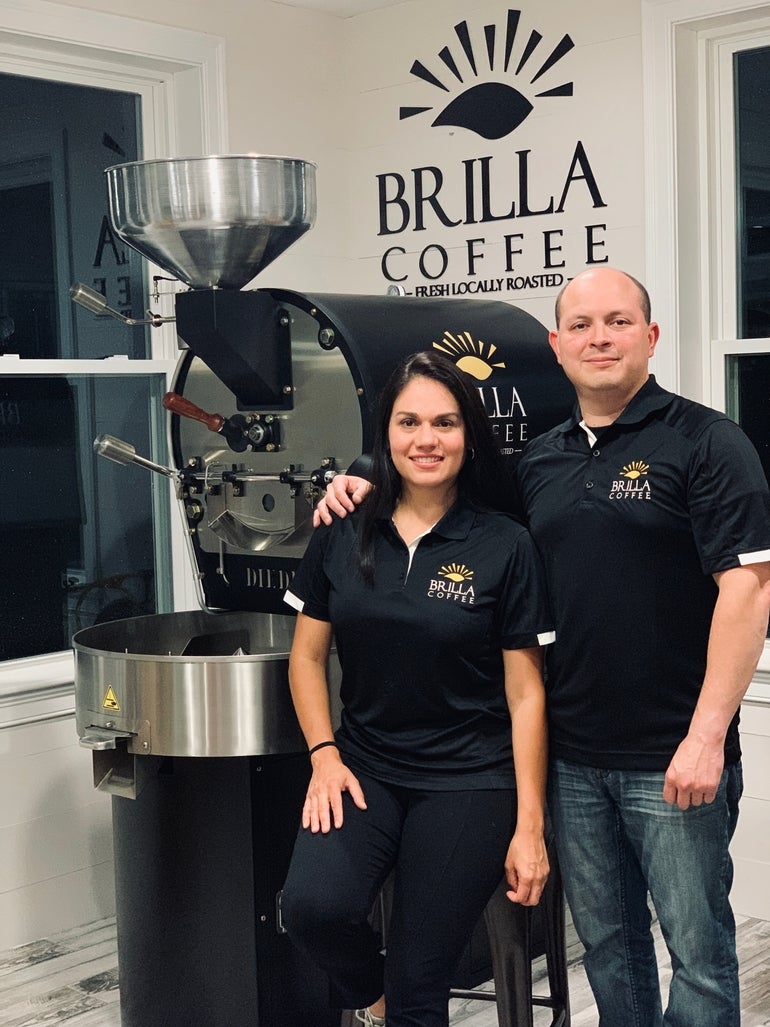 A woman and a man wearing black shirts with the Brilla Coffee logo stand in front of coffee-making equipment.