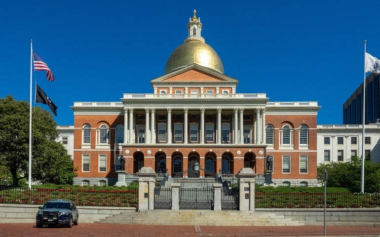 A large brick building with columns and a large gold dome on top sits behind a gate with steps leading up to it.