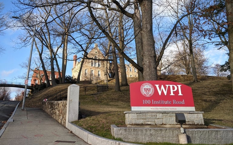 A red sign with "WPI" in white letters in front of an uphill field leading to a brick building.