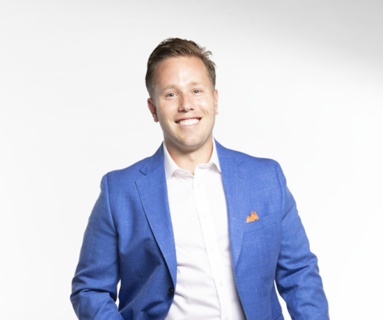 A man with blonde hair wears a white button down and a blue suit jacket.