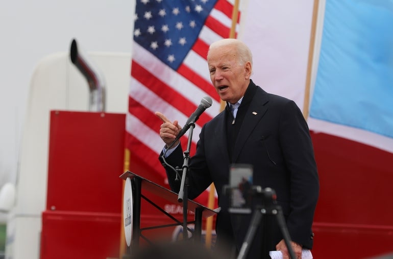 A man with white hair and a black suit jacket speaks at a podium in front of an American Flag.