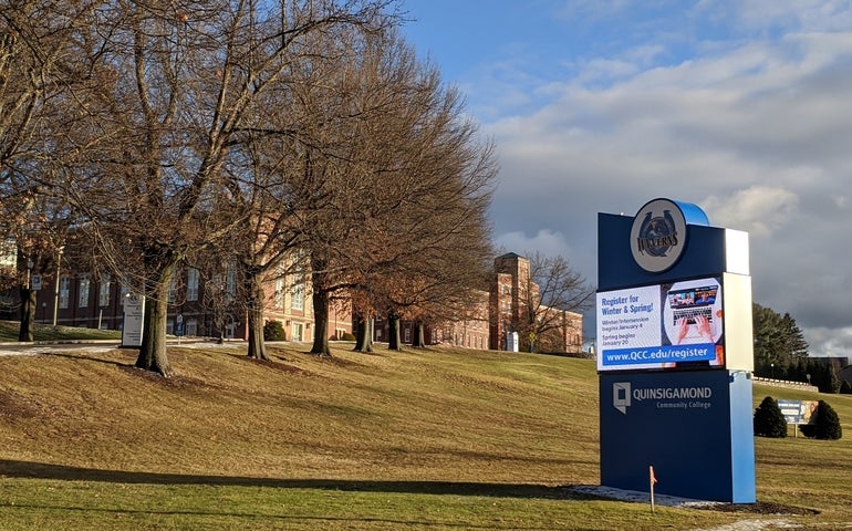 A photo of a sign for QCC sitting on a slightly sloping lawn