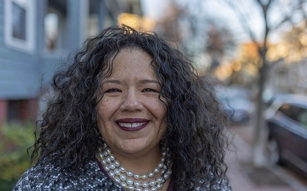 A woman with dark curly hair wears purple lipstick, large pearl necklaces, and a patterned top standing on a neighborhood street.