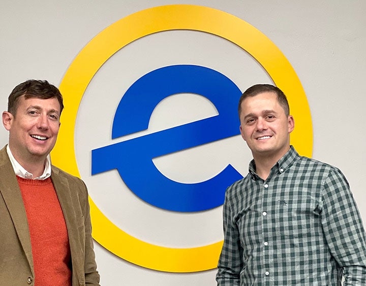 Two men dressed in business casual smile in front of a large blue and yellow logo featuring the letter E
