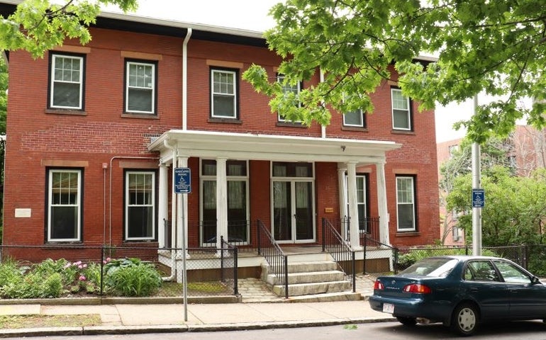 A brick building with a white-columned porch with a garden in front