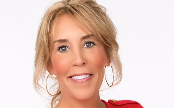 A headshot of a woman with blond hair tied back and bangs wears gold hoop earrings and a red blouse.
