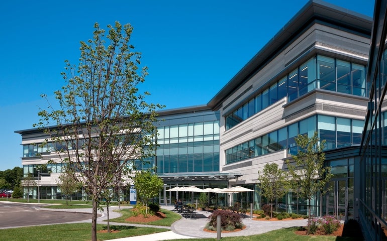 A building with blue tinted windows sits behind a patio with chairs and umbrellas. 