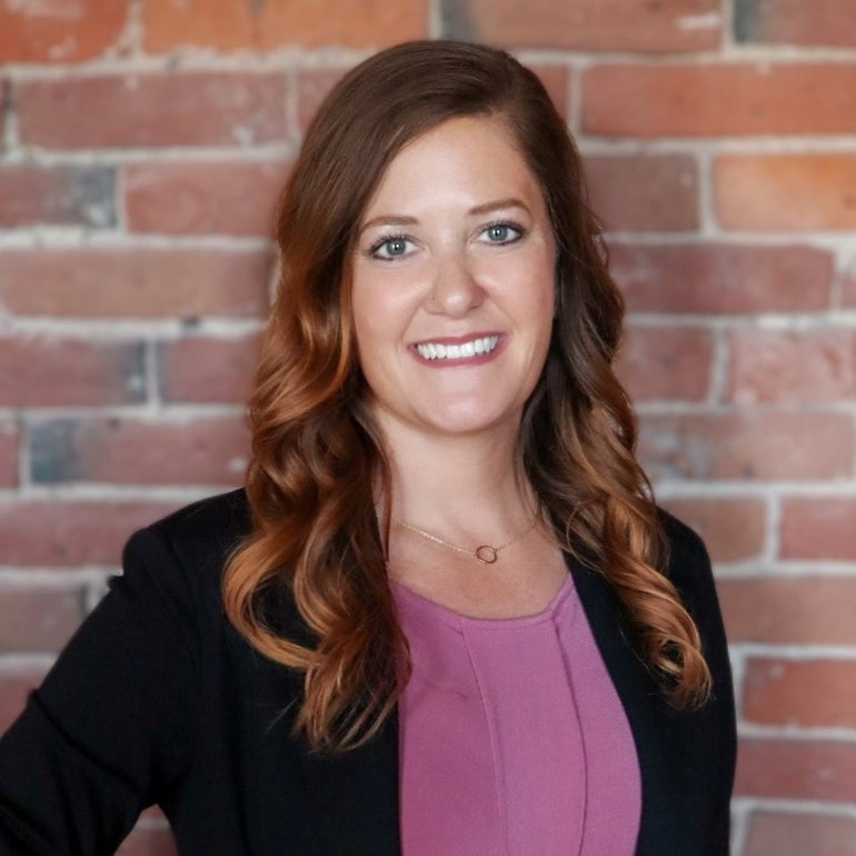 A photo of Julie Bowditch wearing a pink top and black blazer in front of a brick wall.