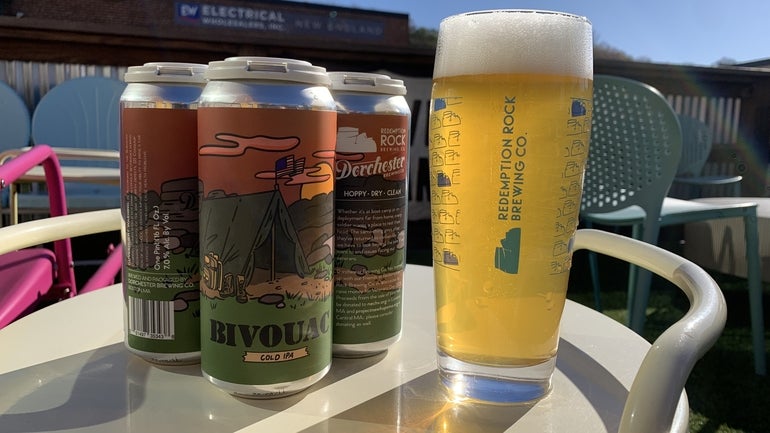 Four canned beers and a glass of beer sit on a table outside