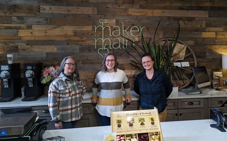 Three women stand behind a counter, in front of a sign that reads Maker to Main