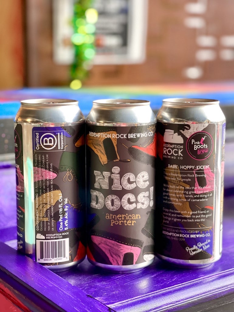 Redemption Rock Brewing Company's Pink Boots 2023 beer is an American Porter called Nice Docs and here cans of it sit on a purple table. 