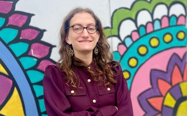 A woman with glasses and brown hair stands in front of a colorful mural.