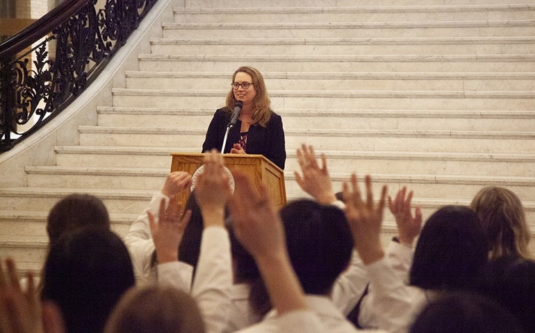 A woman wearing glasses smiles behind a podium on stone steps with a crowd in the foreground.