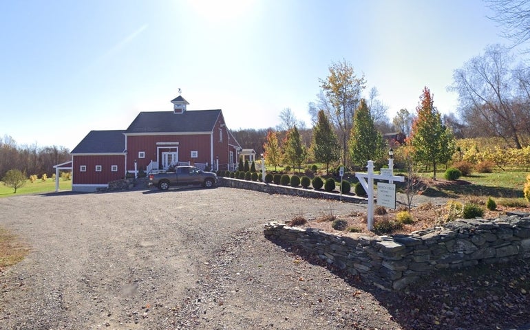 A red barn surrounded by fields and trees with a dirt parking lot and well-kept stone wall
