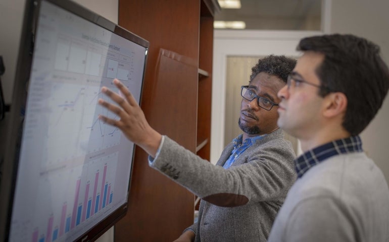 Two men look at a screen filled with graphs