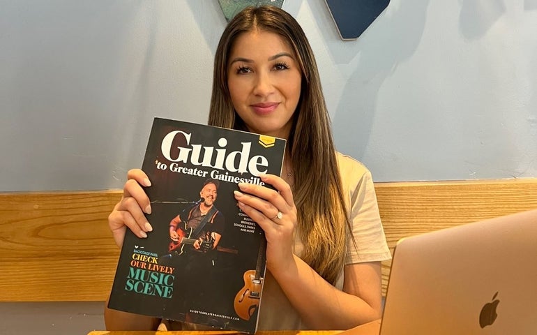 A woman holds a Guide to Greater Gainesville magazine with a Macbook in front of her and the letter W hanging over her head behind her