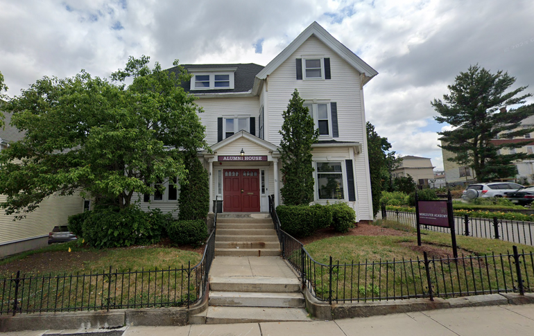 A white three story house with a red sign that says "Alumni House" on it. There's a lawn surrounded by a small iron fence in front of the house. 