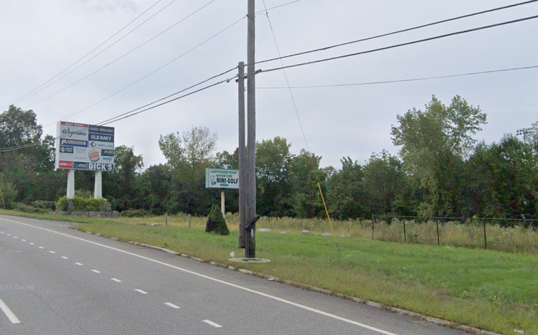 A lot of empty land with short grass in front and longer grass behind it. A small sign for a defunct minigolf course sits in front of a large pylon sign with advertisements on it.