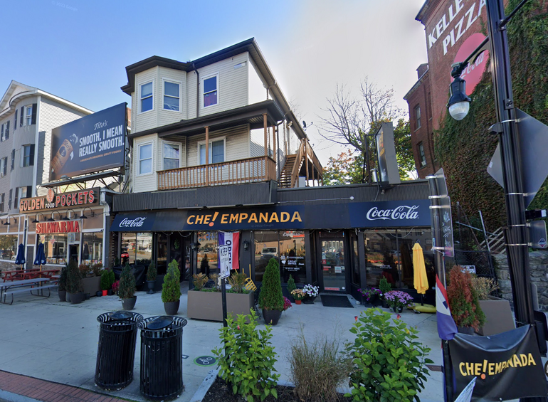 a three story building with a storefront on the bottom and apartments resembling a house on top. Plants and two trash cans sit in front of the property on the sidewalk. 