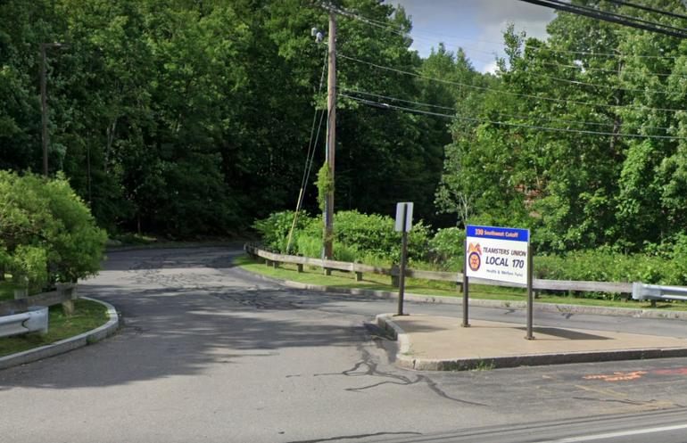 An entrance to a driveway in a wooded area, with a sign on a traffic island in front.