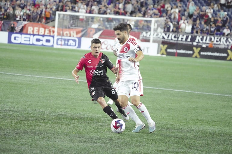 A soccer player dribbles a ball at his feet while a defender chases him.