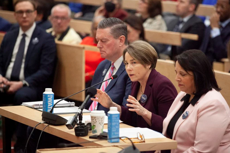 A woman speaks into a microphone at a table, flanked by two other people.