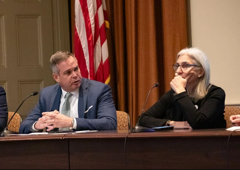 A man and a woman sit at a table with microphones on it