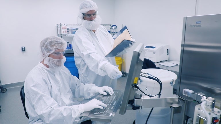 Two workers wearing white lab suits and hair nets covering their faces point at a computer screen.