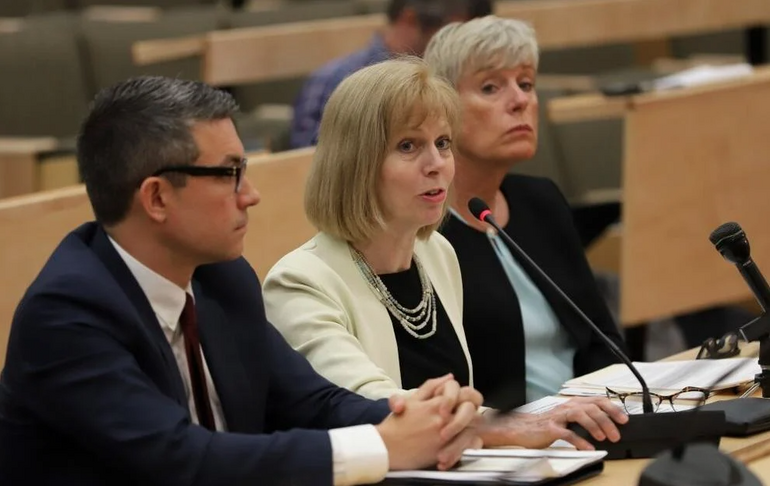 A woman, flanked by a man on her right and a woman on her left, speaks at a hearing while sitting